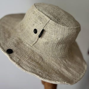 May include: A beige, wide-brimmed hat with a textured weave. The hat features a rounded crown and a wide brim. Two black buttons are visible on the side. The hat is made of a woven material.