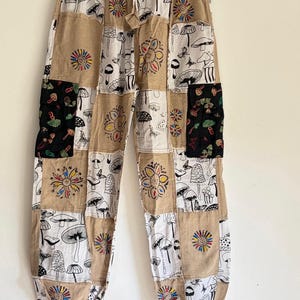 May include: Patchwork trousers featuring a tan and white color scheme with a repeating pattern of black and white mushrooms, butterflies, and colorful sun motifs. The garment includes two black cargo pockets with mushroom prints and an elastic waistband.