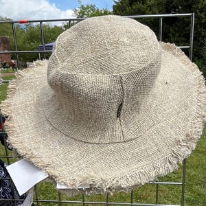 May include: A beige, wide-brimmed sun hat made of woven fabric. The hat features a rounded crown and a frayed edge around the brim. The hat is displayed on a metal grid, with a grassy background.