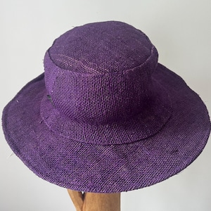 May include: A woven purple sun hat with a wide brim and a rounded crown. The hat is made of a textured material and appears to be handmade. The hat is displayed on a wooden stand against a plain white background.