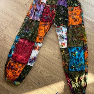 Puede incluir: Pantalones de patchwork con una vibrante mezcla de colores y patrones. Los pantalones presentan una variedad de cuadrados de tela en tonos rojos, naranjas, verdes, azules y morados. La cintura elástica y los puños en los tobillos proporcionan un ajuste cómodo. Una adición única a cualquier armario.