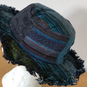 May include: A patchwork bucket hat with a frayed edge. The hat features various fabric patterns, including geometric designs, stripes, and a dark blue colour scheme. The hat is displayed on a white mannequin head.