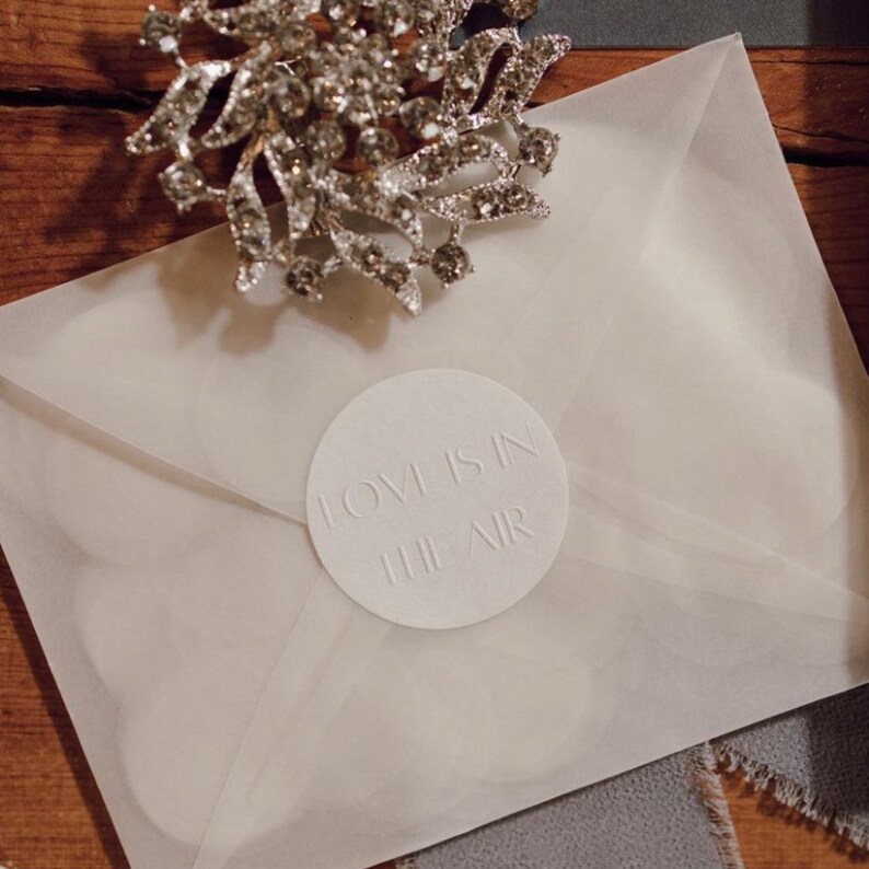 Op de afbeelding: Close-up van een trouwuitnodiging met een witte envelop en een ronde, reli&euml;fsticker met de tekst "LOVE IS IN THE AIR". Een zilveren strass-broche ligt erop. De uitnodiging ligt op een houten oppervlak.