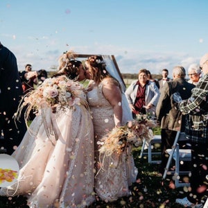 May include: Two brides share a kiss during an outdoor wedding ceremony. They are surrounded by guests and confetti. The brides wear white wedding dresses and hold bouquets of flowers. The sky is blue with a few clouds.
