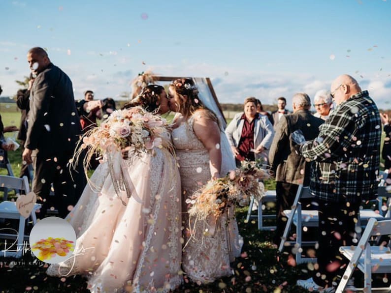 May include: Two brides share a kiss during an outdoor wedding ceremony. They are surrounded by guests and confetti. The brides wear white wedding dresses and hold bouquets of flowers. The sky is blue with a few clouds.