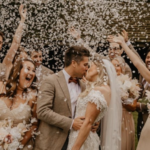 May include: A wedding celebration with a newly married couple kissing, surrounded by friends and family. Confetti is falling from above. The bride wears a white lace gown and veil, while the groom wears a brown suit and bow tie. The wedding party holds bouquets.