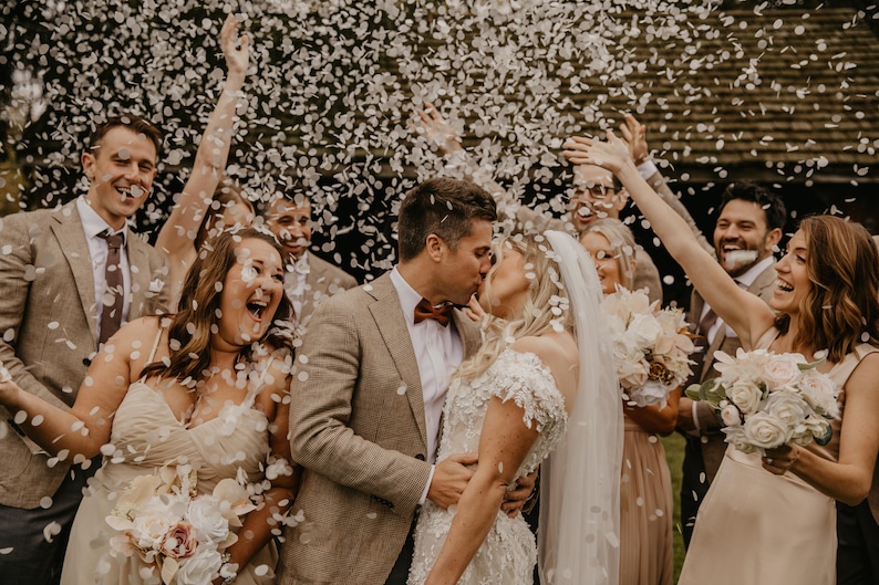 May include: A wedding celebration with a newly married couple kissing, surrounded by friends and family. Confetti is falling from above. The bride wears a white lace gown and veil, while the groom wears a brown suit and bow tie. The wedding party holds bouquets.
