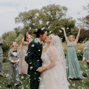 Peut inclure: Sc&egrave;ne de mariage avec un couple s'embrassant, entour&eacute; de confettis. La mari&eacute;e porte une robe blanche et un voile, le mari&eacute; un costume vert fonc&eacute;. Les invit&eacute;s sont en tenue de c&eacute;r&eacute;monie, en ext&eacute;rieur.