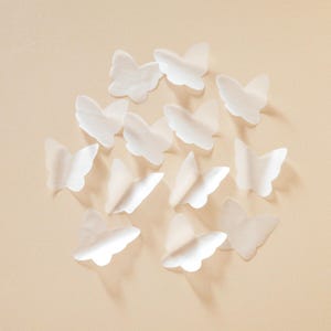 Peut inclure: Plusieurs d&eacute;corations en forme de papillons blancs, en mat&eacute;riau semblable &agrave; du papier, dispos&eacute;es sur un fond beige clair. Les papillons semblent d&eacute;licats et translucides, avec des ombres douces qui ajoutent de la profondeur. Les ailes sont l&eacute;g&egrave;rement incurv&eacute;es.