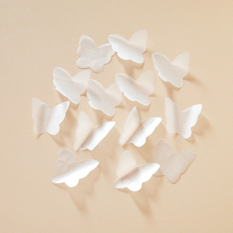 Peut inclure: Plusieurs d&eacute;corations en forme de papillons blancs, en mat&eacute;riau semblable &agrave; du papier, dispos&eacute;es sur un fond beige clair. Les papillons semblent d&eacute;licats et translucides, avec des ombres douces qui ajoutent de la profondeur. Les ailes sont l&eacute;g&egrave;rement incurv&eacute;es.