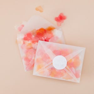 Op de afbeelding: Twee doorschijnende enveloppen gevuld met hartvormige confetti in roze en oranje tinten. E&eacute;n envelop heeft een witte ronde sticker met tekst. Verspreide confetti zweeft rond de enveloppen op een licht perzikkleurige achtergrond.