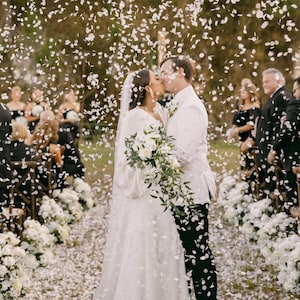 Op de afbeelding: Een bruidspaar kust elkaar tijdens een huwelijksceremonie, omringd door confetti. De bruid draagt een witte jurk en sluier, met een boeket witte bloemen. De bruidegom draagt een wit pak. Gasten zijn op de achtergrond te zien.