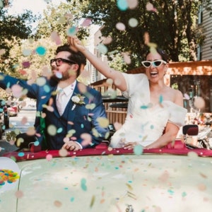 May include: A newly married couple celebrates with confetti in a vintage convertible. The bride wears a white dress and sunglasses, while the groom is in a blue suit. Colorful confetti fills the air, creating a festive atmosphere for a wedding celebration.