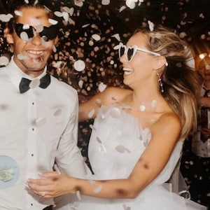 May include: A joyful couple celebrates, surrounded by confetti. The woman wears a white wedding dress and heart-shaped sunglasses. The man wears a white shirt, black bow tie, and sunglasses. The background is blurred, with other people visible.