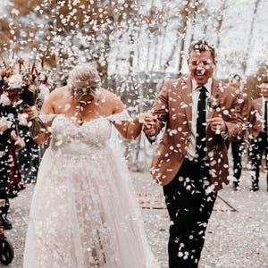 Op de afbeelding: Een pasgetrouwd stel loopt door een confetti regen. De bruid draagt een off-the-shoulder witte trouwjurk met bloemenborduurwerk. De bruidegom draagt een bruine blazer, een zwarte broek en een zwarte stropdas. Andere bruiloftsgasten zijn zichtbaar op de achtergrond.
