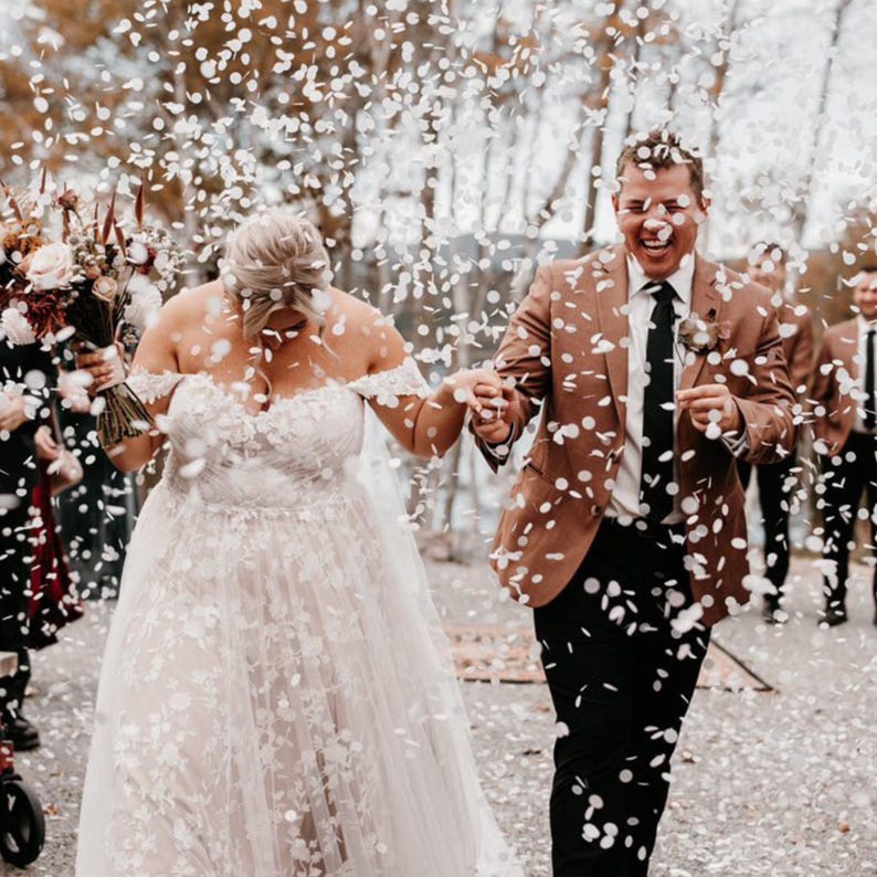Op de afbeelding: Een pasgetrouwd stel loopt door een confetti regen. De bruid draagt een off-the-shoulder witte trouwjurk met bloemenborduurwerk. De bruidegom draagt een bruine blazer, een zwarte broek en een zwarte stropdas. Andere bruiloftsgasten zijn zichtbaar op de achtergrond.