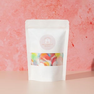 May include: A white pouch with a clear window displaying colorful confetti. The pouch has a circular label with the text "THE CONFETTI FARM". The confetti is a mix of red, yellow, blue, and green colors.