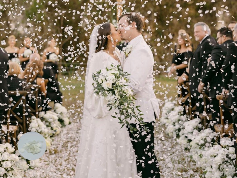 May include: A wedding ceremony with a couple kissing under a shower of white confetti. The bride wears a white gown and veil, holding a bouquet of white flowers. The groom is in a white suit. Guests are in formal attire.