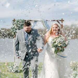 Peut inclure: Un couple souriant, main dans la main, marche sous une pluie de confettis apr&egrave;s leur mariage. La mari&eacute;e porte une robe blanche et tient un bouquet. Le mari&eacute; est en costume gris. Une arche en bois est visible en arri&egrave;re-plan, pr&egrave;s de l'eau.