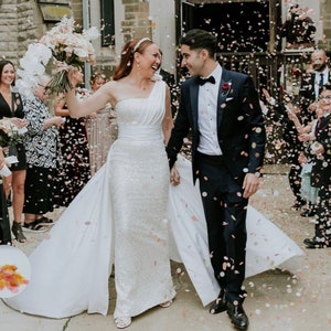 May include: A newly married couple exits a stone building, showered with confetti. The bride wears a white, one-shoulder wedding gown with a long train. The groom is in a navy tuxedo. Guests are visible on either side, celebrating the couple's wedding.