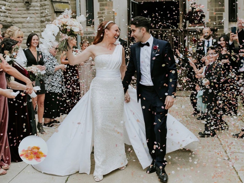 May include: A newly married couple exits a stone building, showered with confetti. The bride wears a white, one-shoulder wedding gown with a long train. The groom is in a navy tuxedo. Guests are visible on either side, celebrating the couple's wedding.