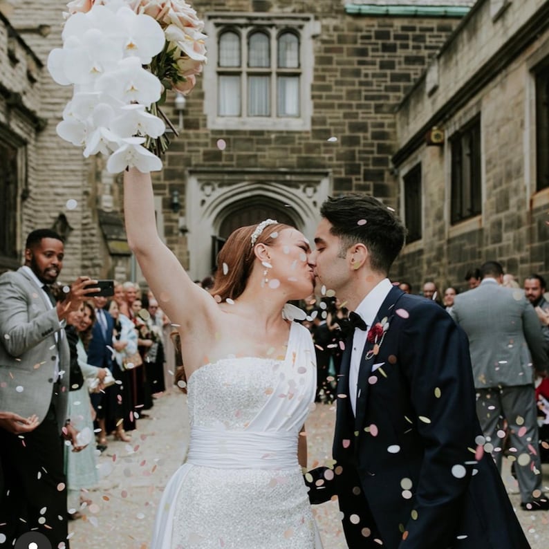 Op de afbeelding: Een pasgetrouwd stel kust elkaar, confetti valt om hen heen. De bruid draagt een witte, off-the-shoulder trouwjurk en houdt een boeket witte orchidee&euml;n en roze rozen vast. De bruidegom draagt een marineblauw smoking. Gasten en een stenen gebouw zijn op de achtergrond te zien.