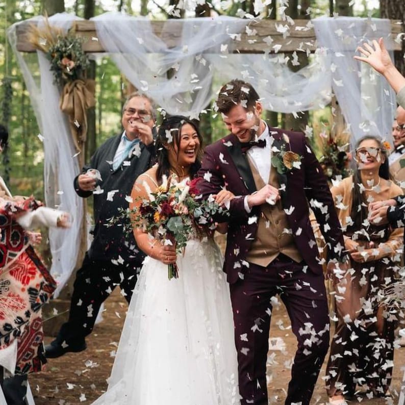 Peut inclure: Une c&eacute;r&eacute;monie de mariage avec une mari&eacute;e en robe blanche et un mari&eacute; en costume bordeaux, entour&eacute;s d'invit&eacute;s. Des confettis blancs tombent d'en haut. La mari&eacute;e tient un bouquet de fleurs. La c&eacute;r&eacute;monie a lieu en plein air sous une arche en bois.