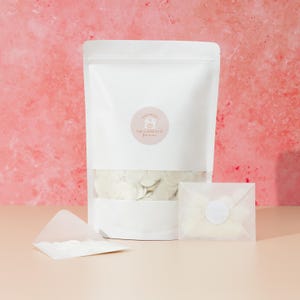 Op de afbeelding: Een witte papieren zak gevuld met witte confetti, een kleine envelop en een doorzichtige envelop met een witte sticker. De zak heeft een ronde sticker met de tekst "THE CONFETTI farm". De achtergrond is roze en perzik.