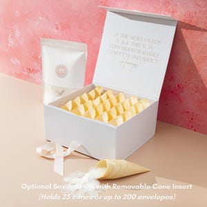 Op de afbeelding: Een witte doos met een magnetische sluiting, gevuld met gele papieren kegels. Een zakje confetti en een kegel met confetti die eruit komt, liggen naast de doos. De tekst op het deksel luidt: "Als de pasgetrouwden deze kant op lopen, gooi dan biologisch afbreekbare confetti en roep Hoera."