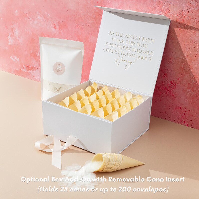 Op de afbeelding: Een witte doos met een magnetische sluiting, gevuld met gele papieren kegels. Een zakje confetti en een kegel met confetti die eruit komt, liggen naast de doos. De tekst op het deksel luidt: "Als de pasgetrouwden deze kant op lopen, gooi dan biologisch afbreekbare confetti en roep Hoera."