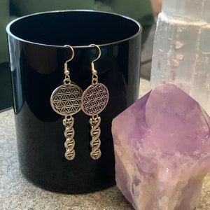 May include: Silver earrings featuring a flower of life design and a twisted, rope-like drop. The earrings are shown on a black surface with a purple crystal.