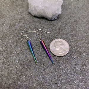 May include: A pair of iridescent silver earrings with a pointed, teardrop shape. The earrings are shown next to a US quarter for size reference.