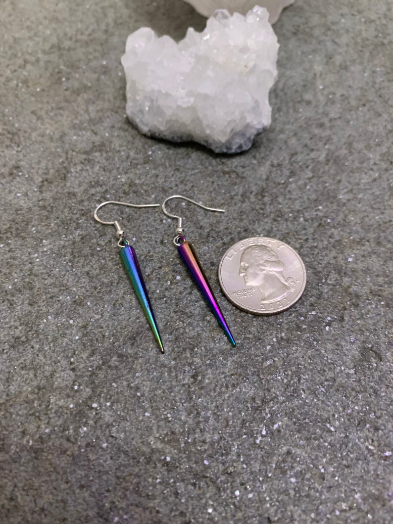 May include: A pair of iridescent silver earrings with a pointed, teardrop shape. The earrings are shown next to a US quarter for size reference.