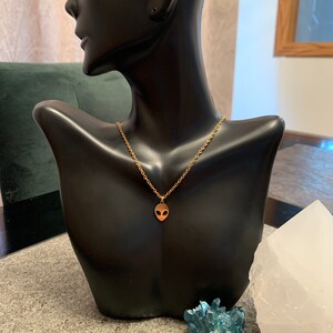 May include: A gold chain necklace with a small alien head pendant. The necklace is displayed on a black mannequin bust.