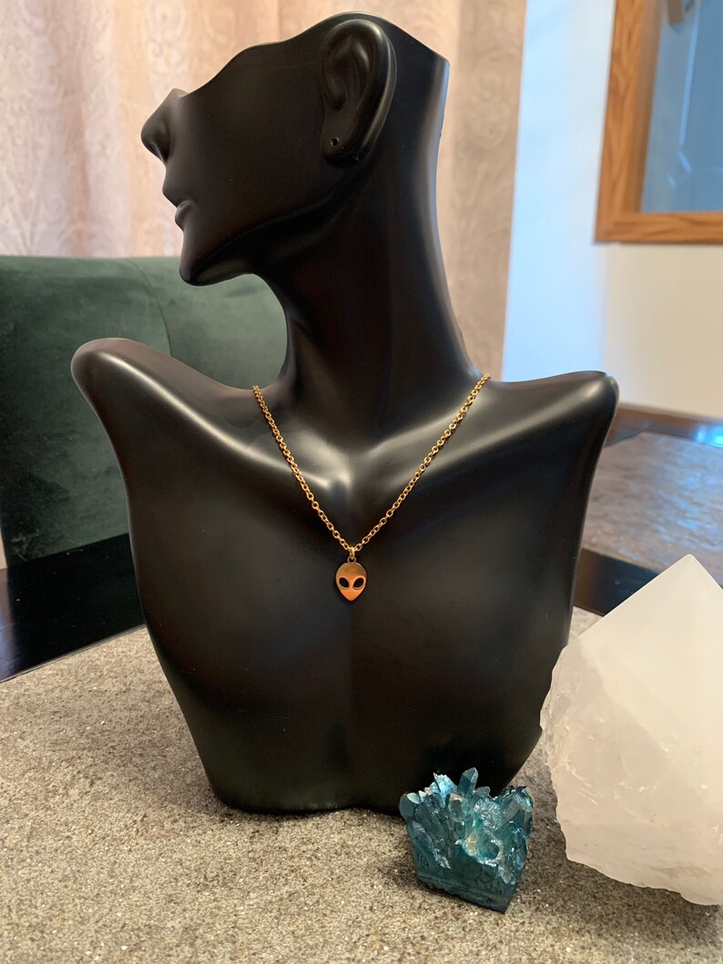 May include: A gold chain necklace with a small alien head pendant. The necklace is displayed on a black mannequin bust.