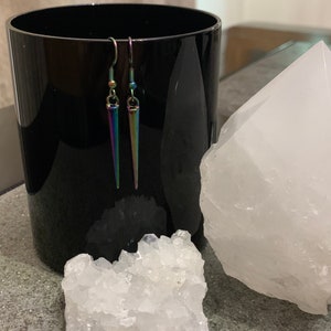 May include: A pair of iridescent silver earrings with a pointed, teardrop shape. The earrings are hanging from a black surface.