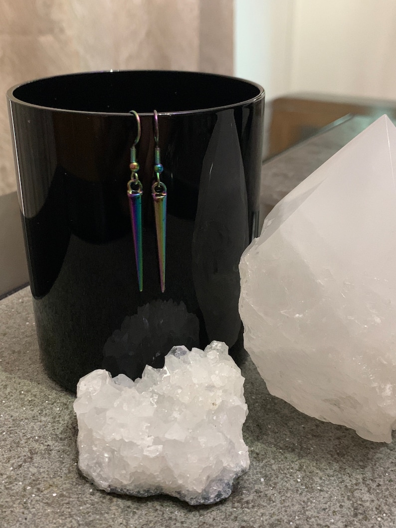 May include: A pair of iridescent silver earrings with a pointed, teardrop shape. The earrings are hanging from a black surface.