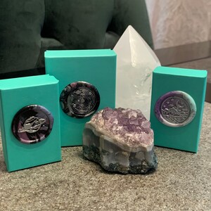 May include: Three teal boxes with different wax seals on top. The seals are black, purple, and silver with different designs. A large amethyst crystal sits in front of the boxes.