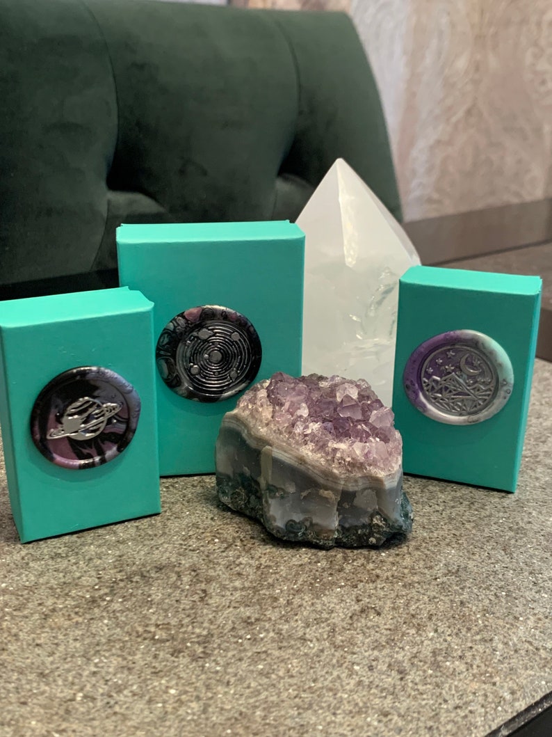 May include: Three teal boxes with different wax seals on top. The seals are black, purple, and silver with different designs. A large amethyst crystal sits in front of the boxes.