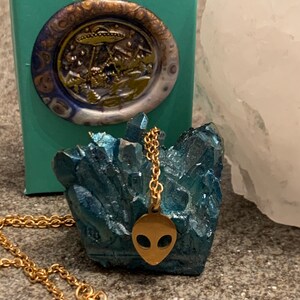 May include: Gold chain necklace with a gold alien head pendant. The pendant is on a blue crystal cluster.