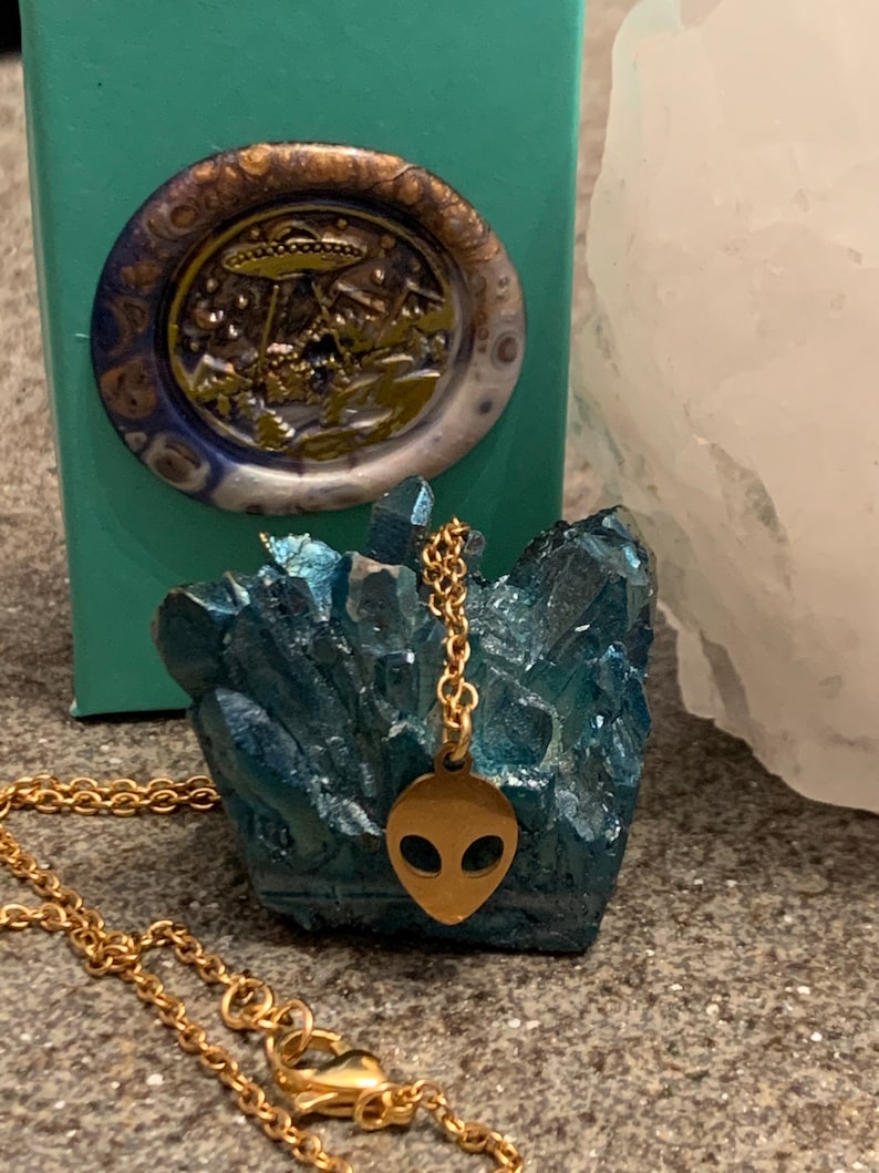 May include: Gold chain necklace with a gold alien head pendant. The pendant is on a blue crystal cluster.
