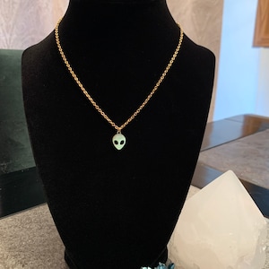 May include: A gold chain necklace with a small green alien pendant. The necklace is displayed on a black velvet stand.