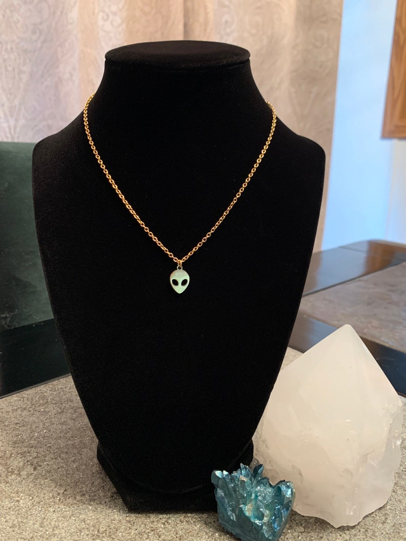 May include: A gold chain necklace with a small green alien pendant. The necklace is displayed on a black velvet stand.