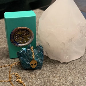 May include: A gold chain necklace with an alien head pendant. The pendant is set on a blue crystal cluster. The necklace is displayed on a turquoise box and a white crystal.