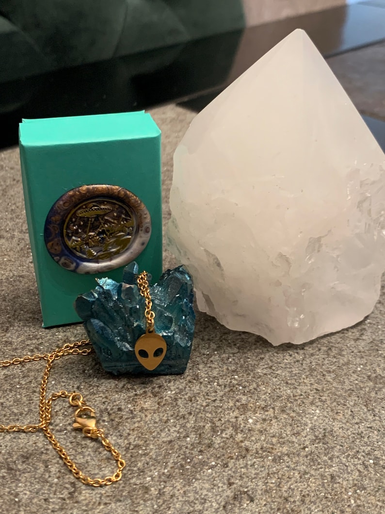 May include: A gold chain necklace with an alien head pendant. The pendant is set on a blue crystal cluster. The necklace is displayed on a turquoise box and a white crystal.