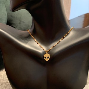 May include: A gold chain necklace with a small alien head pendant. The necklace is on a black mannequin bust.