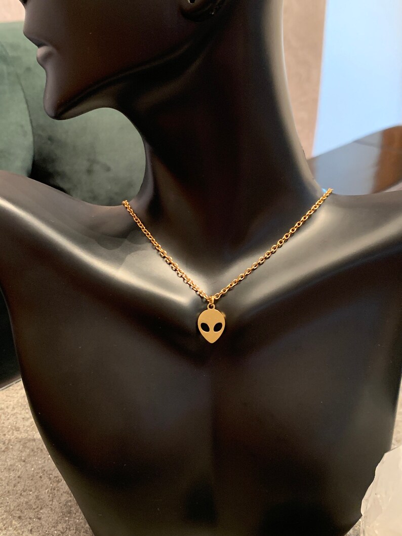 May include: A gold chain necklace with a small alien head pendant. The necklace is on a black mannequin bust.