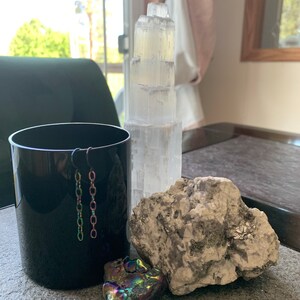 May include: A black ceramic cup with a pair of silver chain earrings hanging from the rim.  A large, rough, gray rock sits next to the cup.  A small, iridescent, multi-colored crystal sits in front of the rock.  A white selenite crystal tower stands behind the cup.