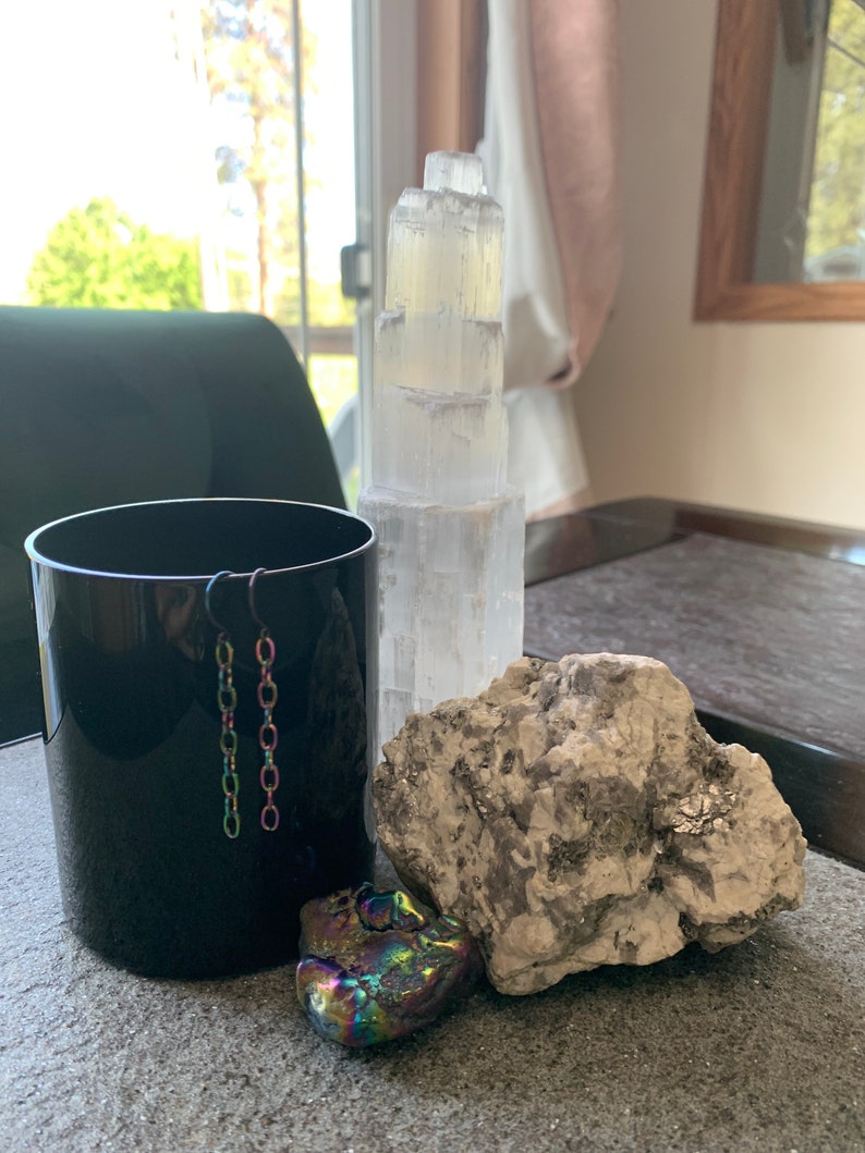 May include: A black ceramic cup with a pair of silver chain earrings hanging from the rim.  A large, rough, gray rock sits next to the cup.  A small, iridescent, multi-colored crystal sits in front of the rock.  A white selenite crystal tower stands behind the cup.