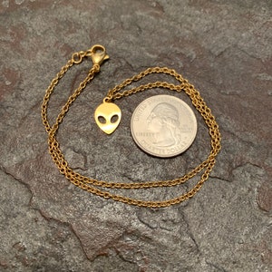 May include: A gold chain necklace with a small alien head pendant. The necklace is laying on a textured surface next to a US quarter.